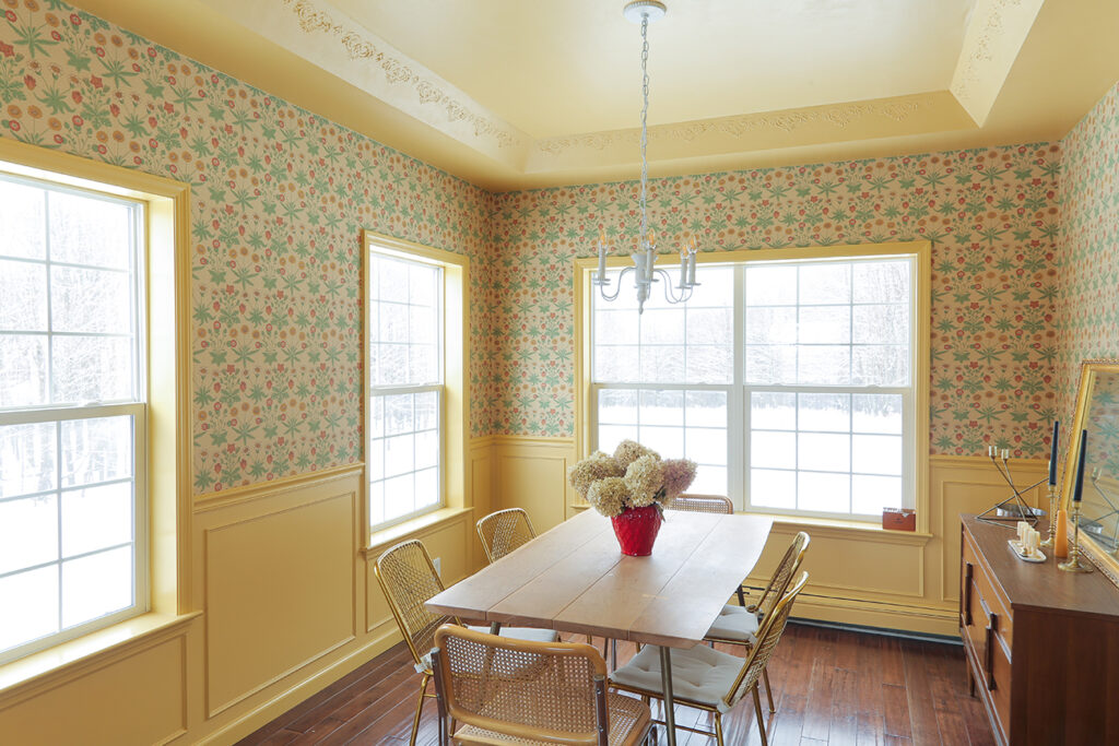 Dining area with brown dining tables and chairs, yellow wainscoting and yellow and blue wall paper with two large windows.