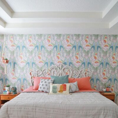 Red and blue wallpaper behind large bed with bright orange and blue throw pillows, two brown end tables on either side.